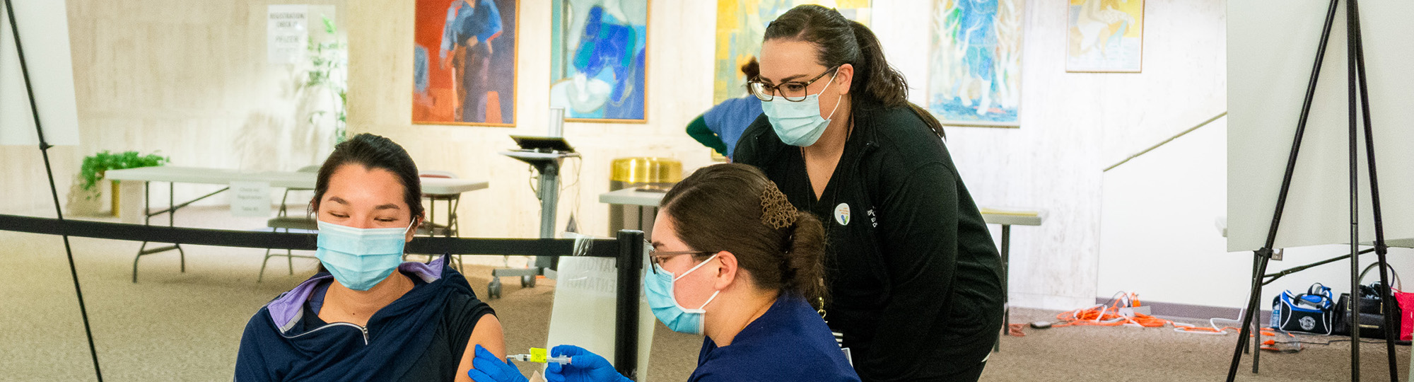 A student administers a vaccine in a patient's upper arm while an instructor looks on.